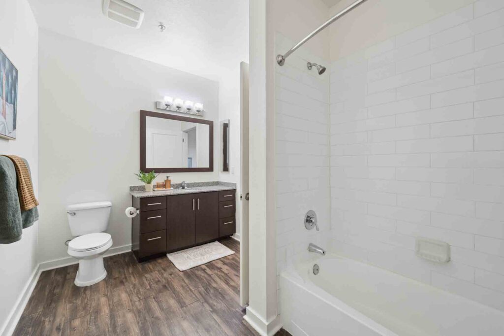 Modern bathroom with white walls, a bathtub with white tile, a toilet, a dark wood vanity with a granite countertop, a large mirror, and wood-look flooring. Towels hang on the wall and a framed picture is displayed.