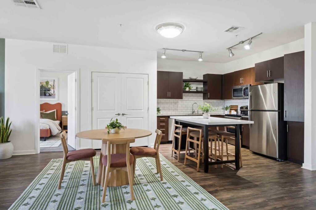 Modern kitchen and dining area with wood flooring, round table, four chairs, island with stools, stainless steel appliances, green plants, and a bedroom visible through an open door.