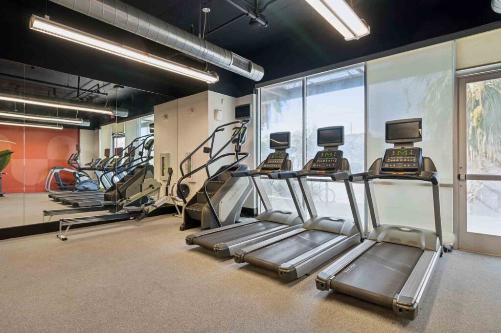 A modern gym with three treadmills, two elliptical machines, and a stair climber facing large windows. The space is well-lit with overhead lights and has a mirrored wall on one side.