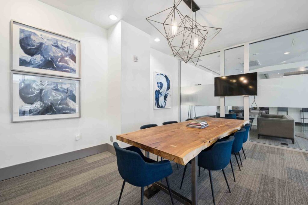 Modern conference room with a wooden table, six blue chairs, abstract art on white walls, a geometric light fixture, a TV mounted on glass wall, and a view into an adjacent office space.