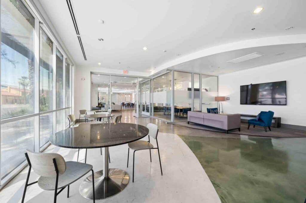 Modern office lounge with round tables, chairs, glass walls, a TV, blue armchair, and gray sofa; large windows let in natural light.