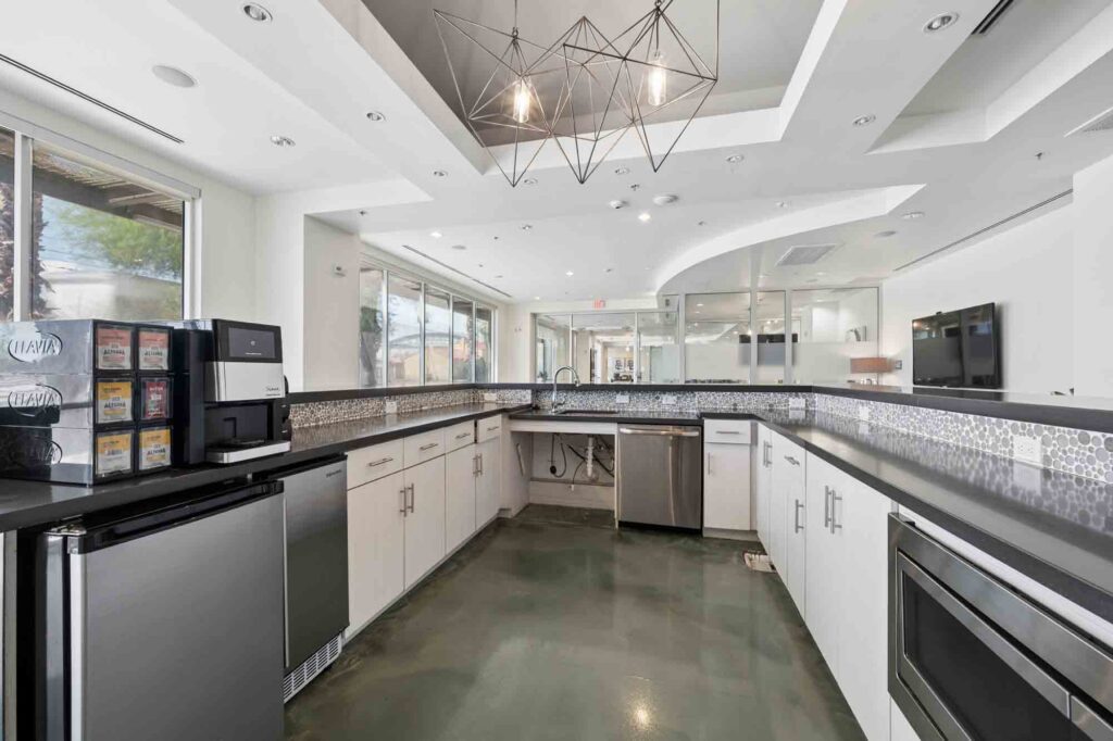 Modern kitchen with white cabinets, stainless steel appliances, a coffee machine, mini fridge, geometric light fixture, and large windows letting in natural light.