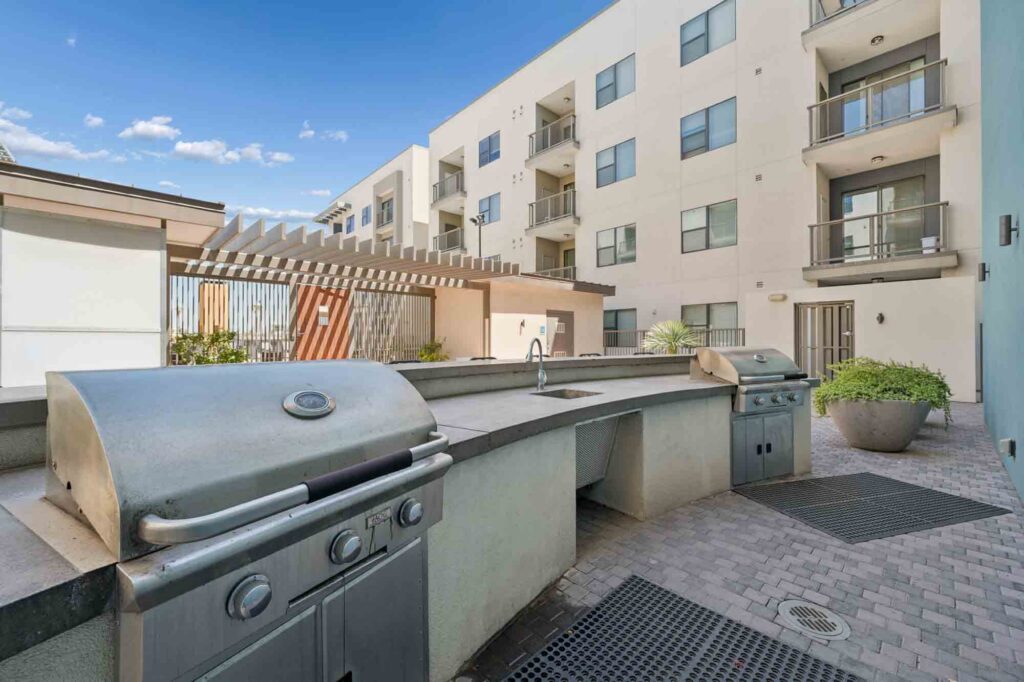 Outdoor patio area with two stainless steel grills, a countertop with a sink, potted plants, and a modern apartment building in the background under a blue sky.