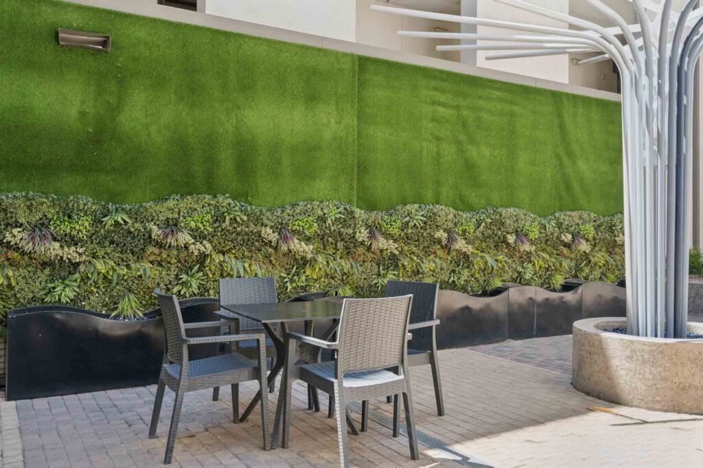 Outdoor seating area with a black table and four woven chairs on a brick patio, in front of a green vertical garden wall with plants. Metal tree-like structures are on the right side.