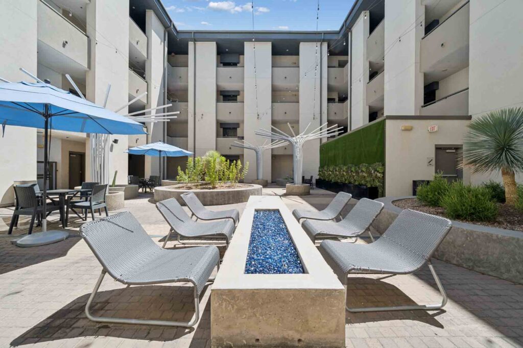 Outdoor courtyard with modern lounge chairs around a rectangular fire pit, blue umbrellas, plants, and a multi-story apartment building in the background under a blue sky.