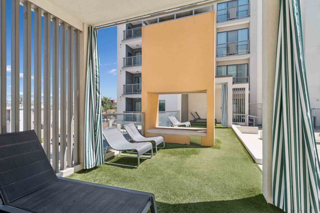Outdoor lounge area with artificial grass, striped curtains, lounge chairs, and a large orange structure with a rectangular opening, set beside a modern apartment building.