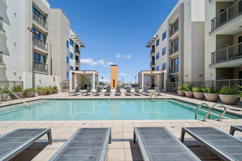 Modern apartment complex courtyard with a rectangular swimming pool, sun loungers, cabanas, potted plants, and three-story buildings on both sides under a clear, blue sky.