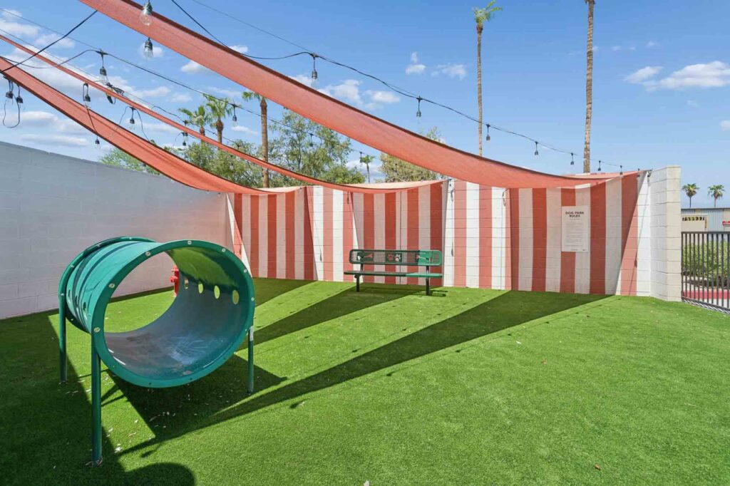 A dog park with artificial grass, a green tunnel, a green bench, red-striped shade sails overhead, string lights, and palm trees visible in the background under a blue sky.