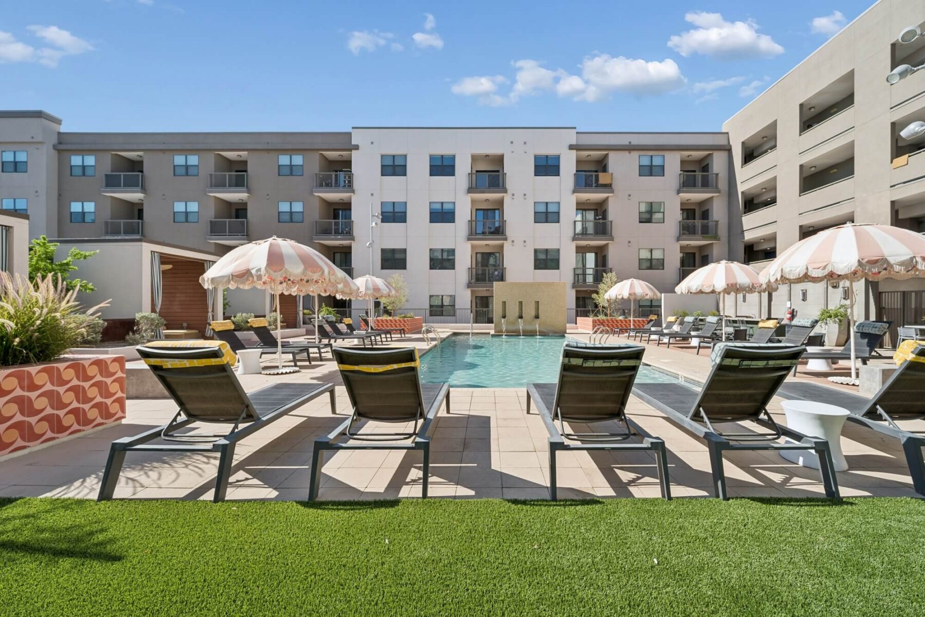 Pool area with lounge chairs, striped umbrellas, and a modern apartment building in the background under a blue sky with scattered clouds.