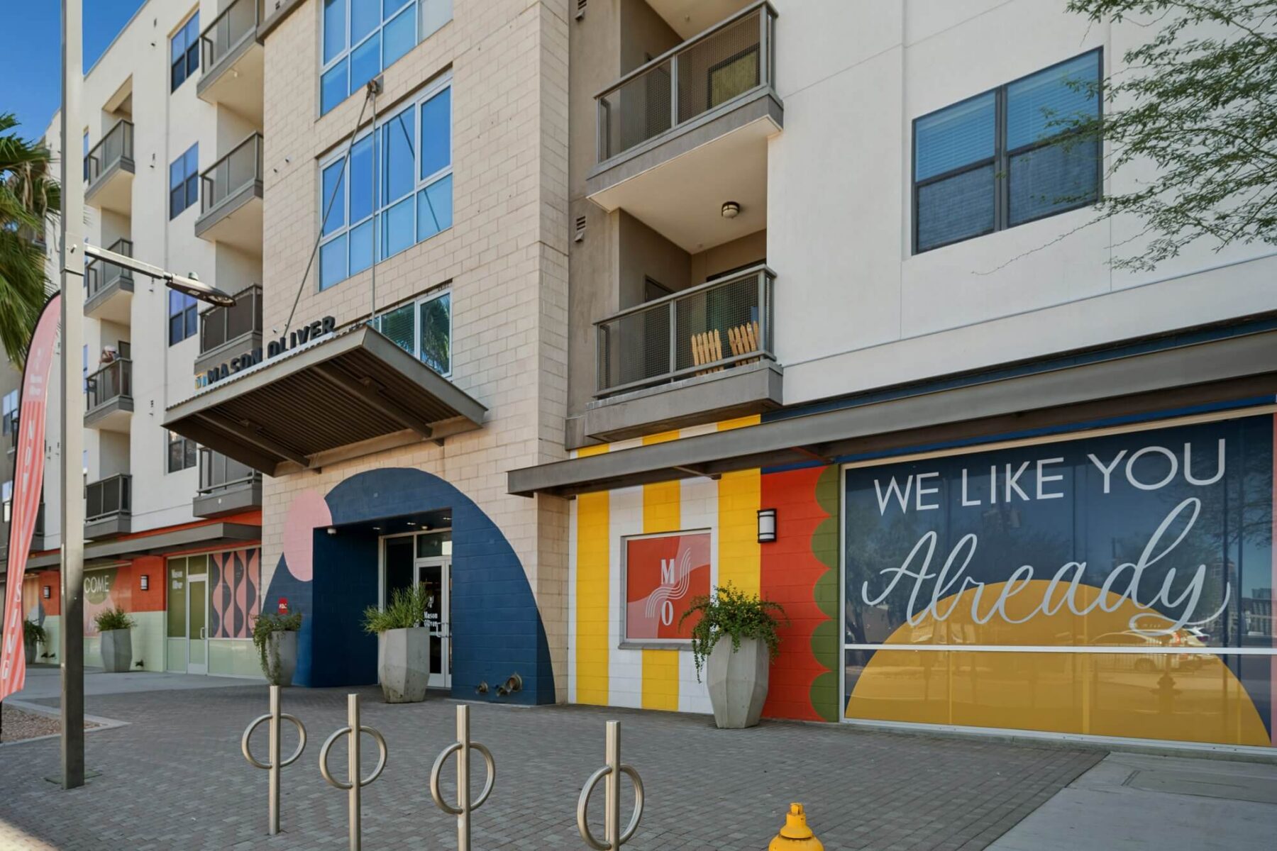 Street view of a modern apartment building with colorful murals and large windows. A sign on the wall reads WE LIKE YOU Already. There are potted plants, a bike rack, and a sidewalk in front.